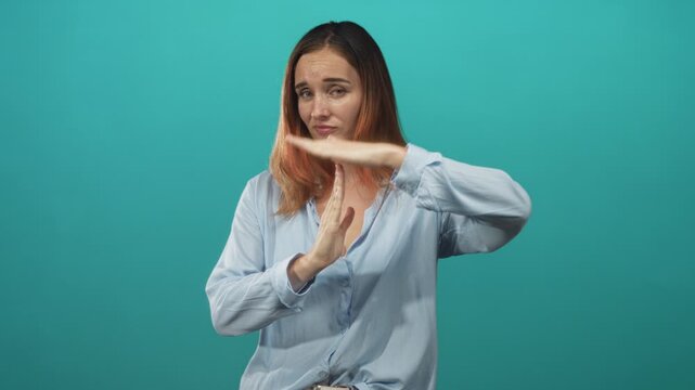 Woman in light blue shirt makes time out gesture with hands in studio teal backdrop, palm horizontal above vertical hand signaling stop; refusal.