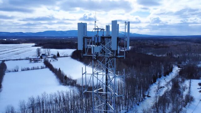 Aerial drone view of a tall relay antenna rising over snowy farmland, bare trees, and distant mountains, showcasing rural telecommunications, winter landscape, and remote connectivity.