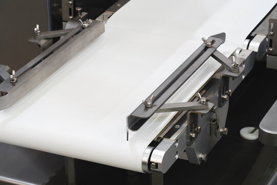 Close-up of industrial conveyor belt with metal guides and rollers. Smooth white belt moves steadily through machinery. Metal parts gleam under bright light