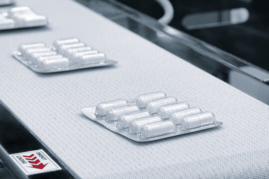 Blister packs move along a factory conveyor belt. White tablets sit neatly in plastic packaging. Industrial setting shows pharmaceutical production. Image fits medical