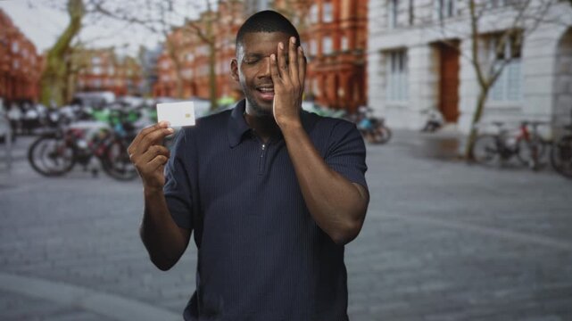Man holding creditcard, hand covering eye on street outside brick buildings and parked bicycles; frustration billing problem.