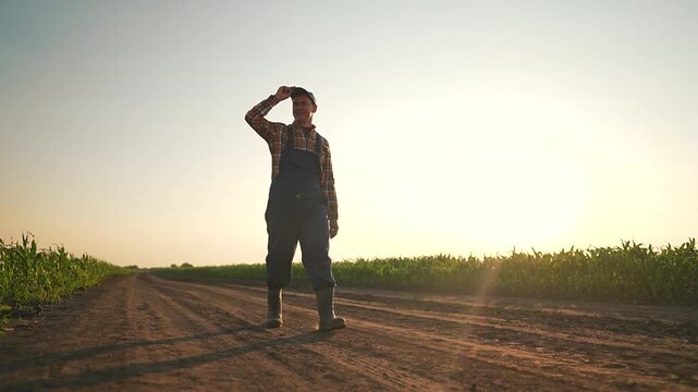 Farmer tips hat on empty road under soft sunset sky. Road cuts through green corn, farmer stands still waving hat. Corn surrounds farmer, quiet road waits, farmer lifts hat proudly facing corn path.