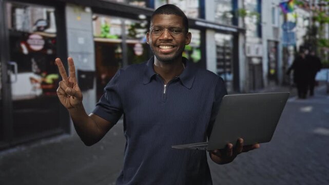Man holding laptop, flashing two finger peace sign with right hand and smiling with closed eyes on a busy city street lined with shops; joy connection.