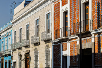 Obraz premium Historic street lined with colorful colonial facades featuring ornate wrought iron balconies and traditional Talavera tiles under bright blue sky in Puebla, Mexico