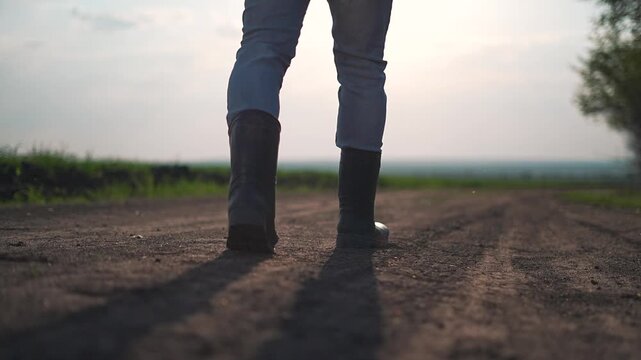 Man in boots steps forward on rural dirt road. Path cuts through green outdoor field. Dirt marks each step as man keeps walking. Motion repeats with boots rising from dust in peaceful outdoor setting.
