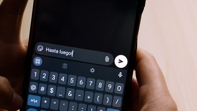 Close-up of hands holding a smartphone while typing "Hasta luego!" (Goodbye!) in a messaging app.