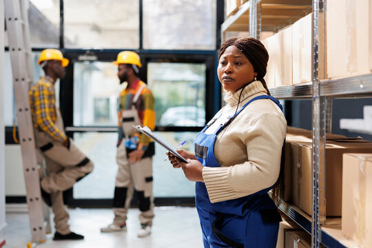 African american warehouse woman operator holding clipboard and standing near carboard boxes shelf. Storehouse supervisor managing merchandise maintenance and dispatching