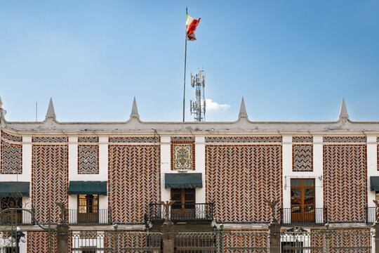 Detailed frontal view of historic Episcopal Palace facade featuring ornate Talavera tiles with geometric patterns, central decorative panel and Mexican flag waving above in Puebla, Mexico