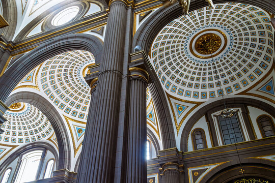 Diagonal low angle view of towering fluted column and ornate coffered ceilings with blue and gold patterns inside cathedral, illuminated by sunlight creating lens flare in Puebla, Mexico