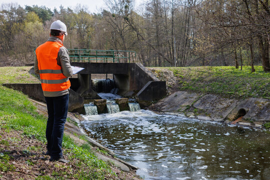 Environmental engineer inspecting a river dam and water level control system for ecological monitoring. Ecologist in safety vest assessing water quality and flow at a small hydraulic dam in the forest