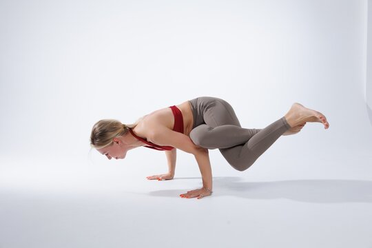 Athletic woman performing an advanced yoga inversion pose in a minimalist studio setting. She is balancing on her forearms with legs extended in a split position, demonstrating strength, flexibility, 