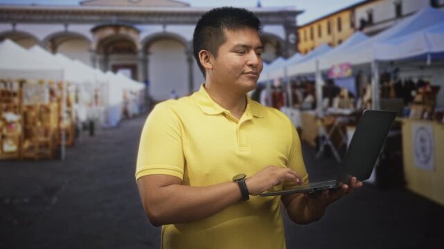 Man wearing yellow polo shirt holding a laptop and typing on keyboard among outdoor market stalls on a street plaza; focused productivity.