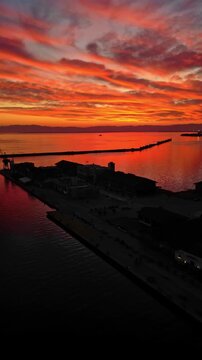 Spectacular vertical drone footage at the Port of Thessaloniki during a vibrant red and orange sunset. The dark silhouettes of the docks, historic buildings, and people contrast beautifully with the d