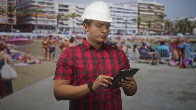 Man architect wearing a hardhat tapping a tablet while holding it on a busy beach promenade; focused planning.
