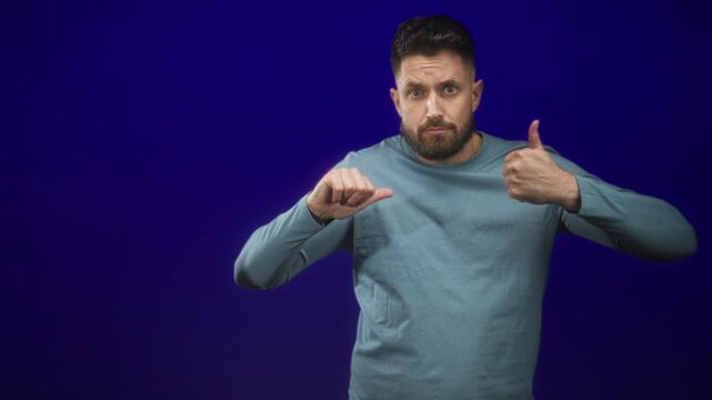 Bearded man shows simultaneous thumbs up and thumbs down with both hands in studio against blue backdrop; ambivalence.