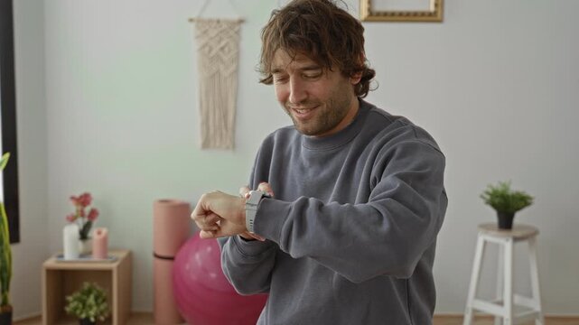 Man pressing stopwatch on wrist in yoga studio with pink exercise ball, mat, plants and macrame wall hanging; focused wellness routine.