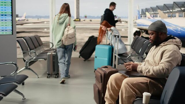 African american connected traveler in busy airport terminal lounge, typing on laptop and waiting for boarding flight. Modern digital nomad job and flexible career with travel lifestyle.