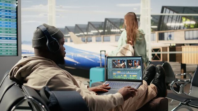 Digital nomad videographer on laptop in airport terminal lounge, cutting and editing video footage for web content while waiting for flight departure. Location independent nomadic lifestyle.