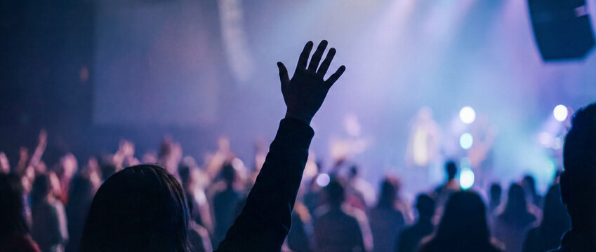 Silhouette of a raised arm and hand in a large crowd at a live music concert or worship service with atmospheric blue stage lights
