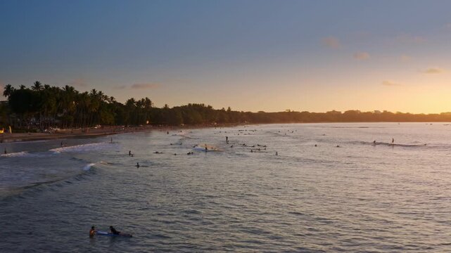 Aerial Forward Tracking Shot of Surfers at Sunset on Tamarindo Beach Costa Rica. The golden hour light illuminates the tropical coastline and palm-fringed shore in a serene Costa Rican paradise.
