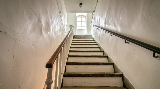 Ascending a staircase with worn white walls and handrails, light from a window at the top