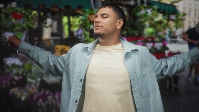 Young hispanic man holding and fanning us dollars with one hand, head tilted and eyes closed, standing in front of a street flower market stall; confidence.