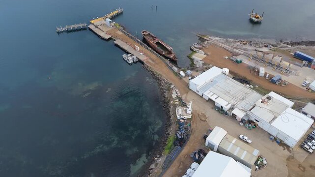 Drone flight over a maritime industrial site toward a massive rusted shipwreck.