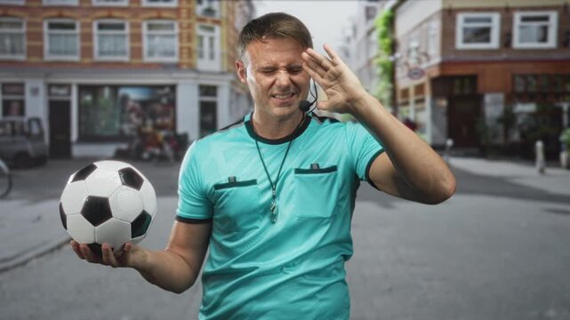 Man referee in teal shirt holding soccer ball and whistle, headset visible, hand to face gesture amid urban street storefronts; tension frustration.