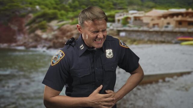 Police officer man clutching stomach, grimacing with stomachache at rocky beach cove with badge visible; pain emergency.