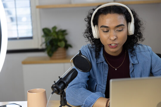 Non-binary person wearing denim jacket and headphones speaking into mic using laptop in home studio
