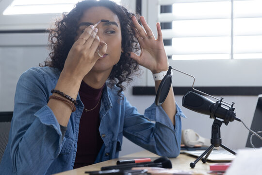 Non-binary adult applying eyebrow makeup with spoolie at home studio desk in denim with microphone