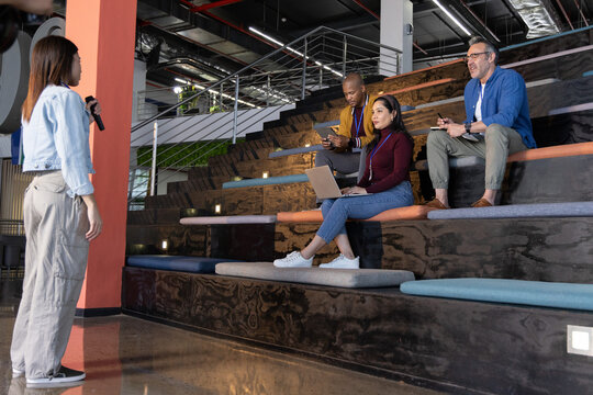 Diverse coworkers listening as Asian presenter on left holding microphone laptop notes, copy space