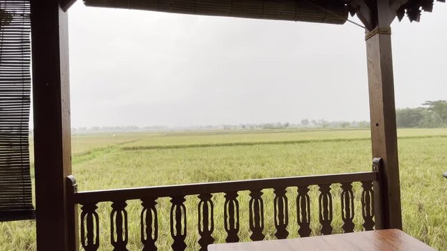 Wide view of rainy farmland from a rustic veranda with wooden posts, railing, and table in the foreground.