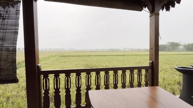 Wide view of rainy farmland from a rustic veranda with wooden posts, railing, and table in the foreground.