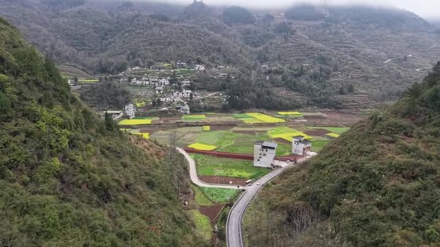 A panoramic drone shot of a remote Chinese village tucked away in a lush mountain valley, surrounded by vibrant golden rapeseed terraces under a hazy spring sky.
