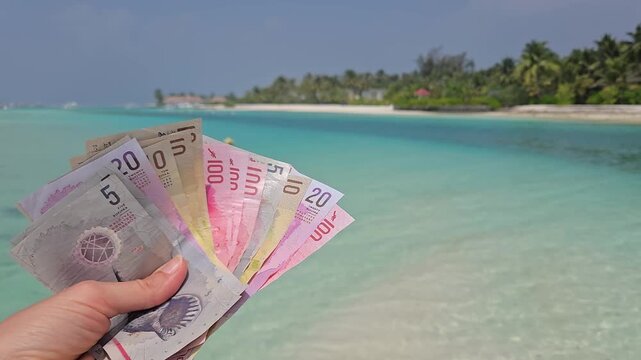Stack of different denominations of Banknotes of Maldives, Maldivian rufiyaa in hand with maldivian island and ocean in background. Expensive travel concept