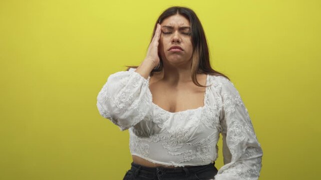 Woman wearing white embroidered top holding hand to temple for headache and closing eyes in a bright yellow studio; pain.