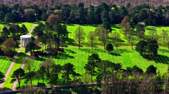Aerial sideways drone reveals Normandy American Cemetery with vivid spring lawn, tree-shadowed white cross rows, and rotunda chapel surrounded by forest in Normandy, France
