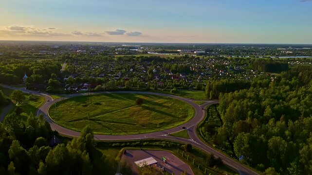 Drone parallax orbit at golden sunset reveals large traffic roundabout surrounded by lush trees with residential suburbs, church spire and river stretching across flat Jelgava cityscape in Latvia
