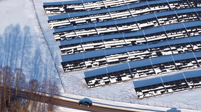 Aerial drone flyover of large solar farm in winter Latvia with rows of snow-covered photovoltaic panels, snowy fields, road and bare trees alongside