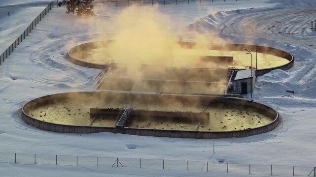 Aerial view of steaming wastewater treatment basins in a snowy industrial landscape, with warm vapor rising over circular tanks during freezing winter conditions.