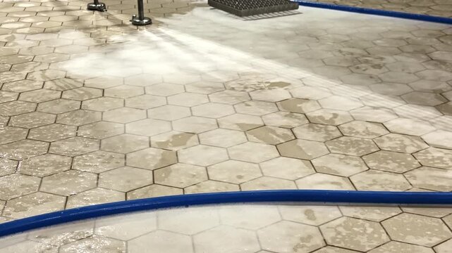 Tilt-up shot of hexagonal tiled industrial facility floor being cleaned with high-pressure water spray showing drains and stainless steel equipment