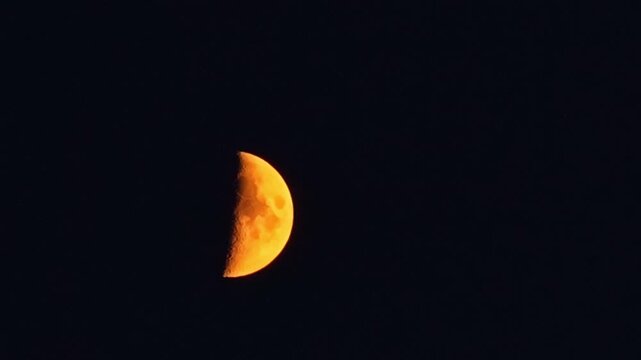 Telephoto time-lapse of vivid orange amber half moon rising against pure black night sky with visible crater surface detail and no foreground
