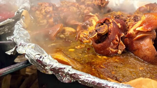 Huge outdoor griddle basins steam with red sausages, pig trotters, chicken and braised meats cooking at busy Latvian winter street food market