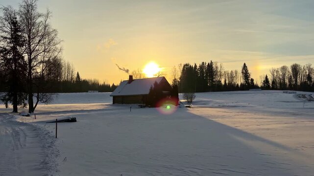 Winter sunset over a snow-covered field with a small countryside house, chimney smoke, and forest edge, creating a calm rural landscape scene in golden cold light.