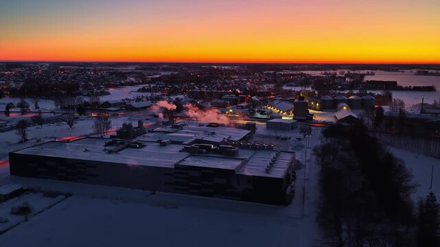 Aerial winter twilight over a snowy town and industrial area with glowing horizon, lights, and steam plumes, capturing cold evening atmosphere and urban-rural landscape.