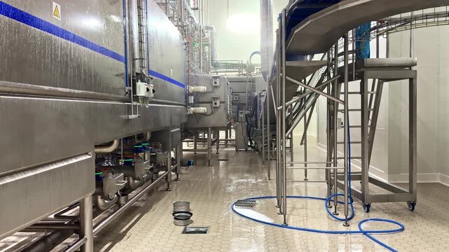 Wet washdown inside a modern food processing factory with stainless steel machinery, conveyor structures, steam spray, and hoses during industrial sanitation cleaning.