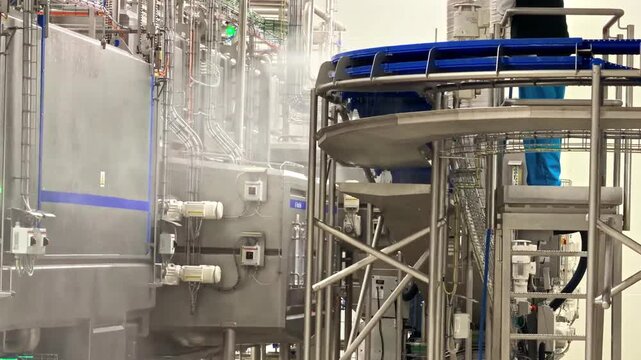 Worker steam washing large stainless steel processing tanks with pipes and conveyors inside modern dairy production facility with blue industrial equipment