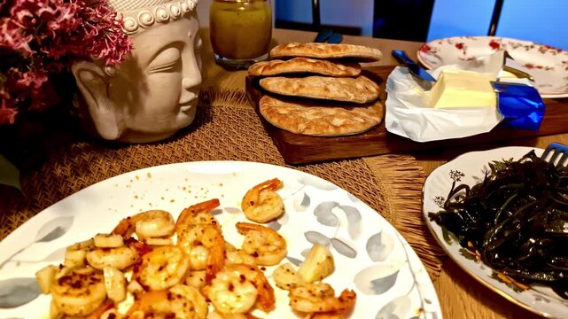 Home dining table with garlic shrimp, seaweed salad, crusty bread and butter, styled with a Buddha decor piece&mdash;cozy homemade seafood meal in warm indoor light.