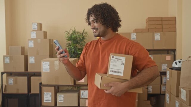 Man holding smartphone and cardboard boxes while smiling and checking messages in a building; small business pride.